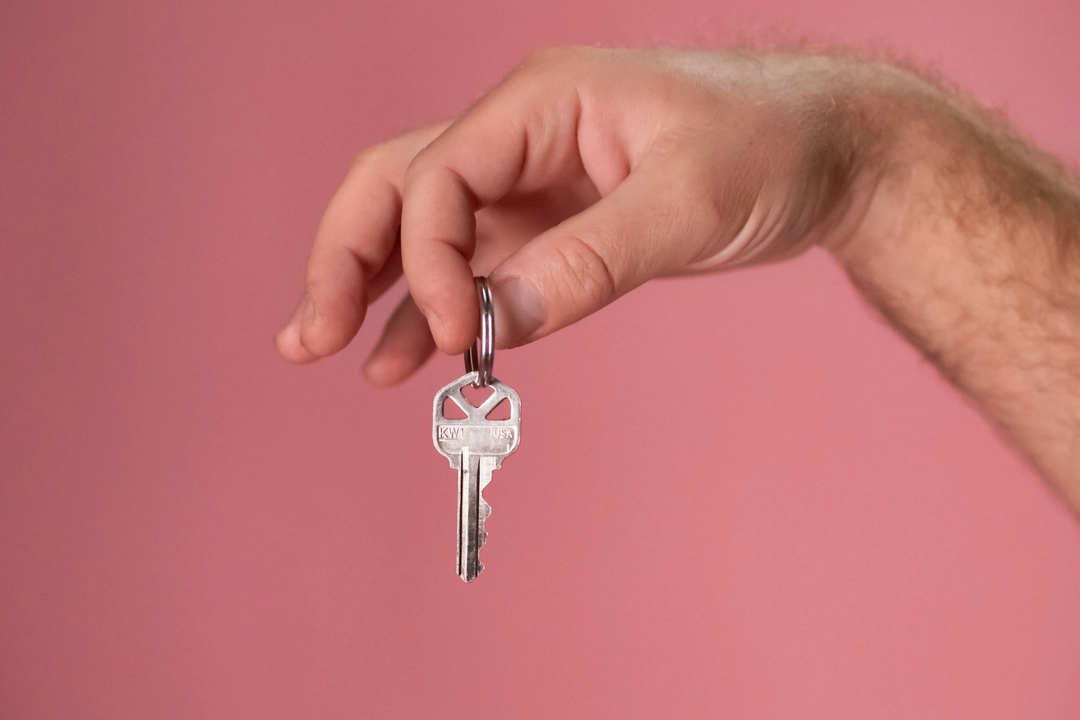 A close-up image of a person's right hand, with light skin tone, holding a silver key with a bow labeled 'KW1'. The key is displayed prominently against a plain pink background, filling the central area of the frame. This visual metaphorically represents the concept of security and access, akin to the process of learning how to create and use SSH keys for secure remote access.