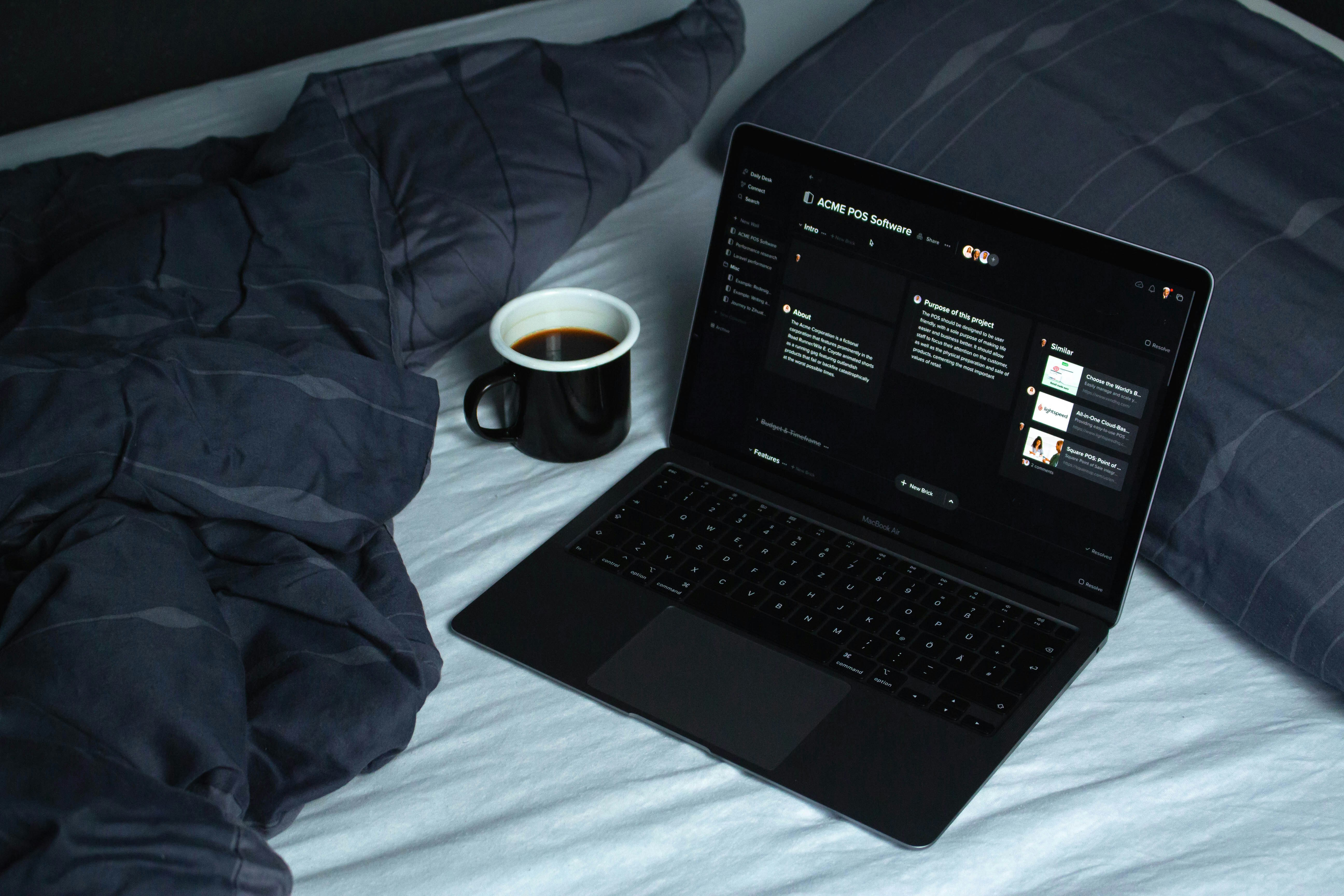 A laptop displaying a project on ACME POS Software sits on a bed next to a cup of black coffee. This cozy setup illustrates a casual working environment, ideal for those researching "Regex performance optimization and security best practices" in a relaxed setting. The dark-themed screen contrasts with the dark bedding, creating a focused ambiance for programming or studying advanced coding techniques.